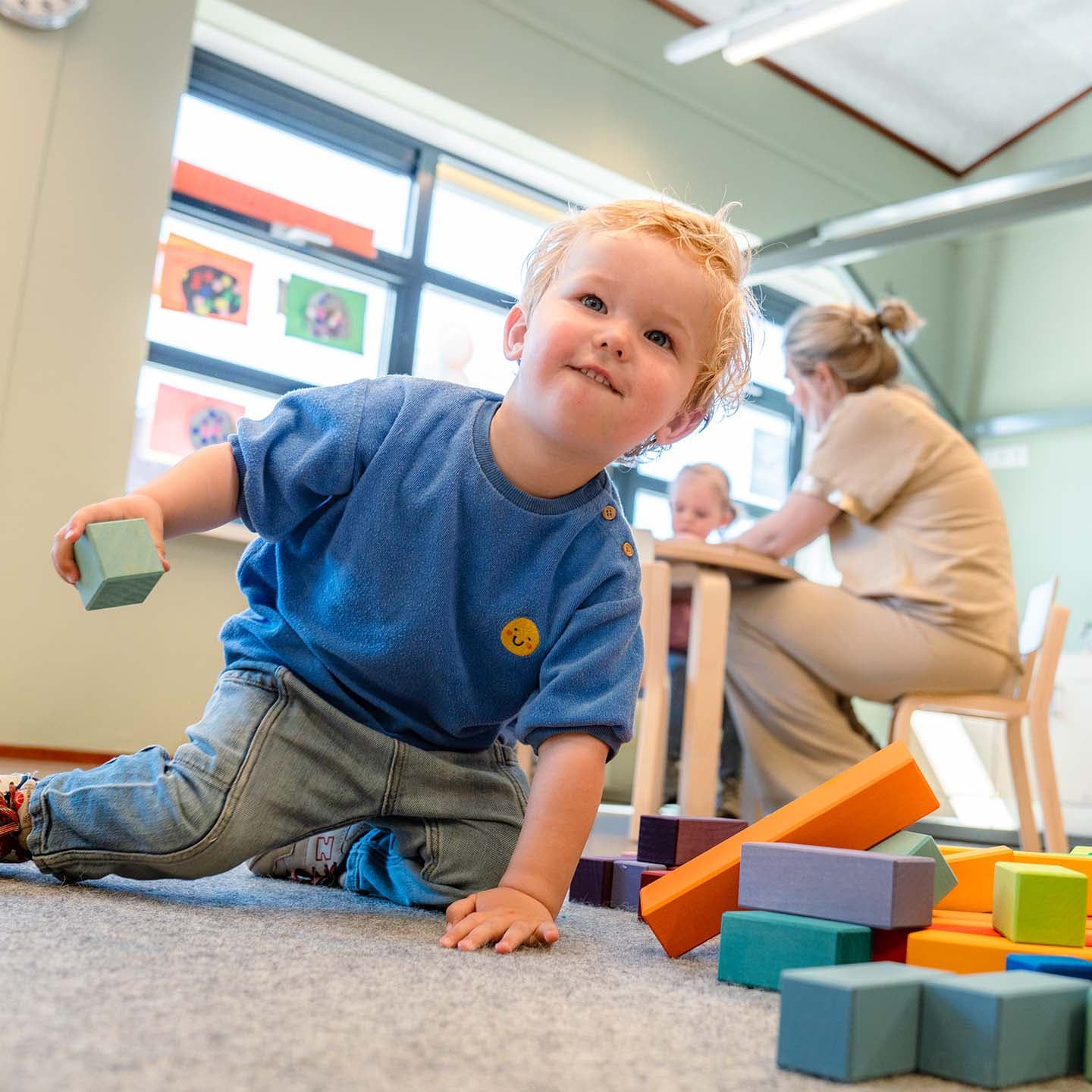 Een jong kind in een blauw shirt speelt met kleurrijke blokken op de grond, terwijl een pedagogisch professional en een ander kind op de achtergrond aan een tafel zitten.