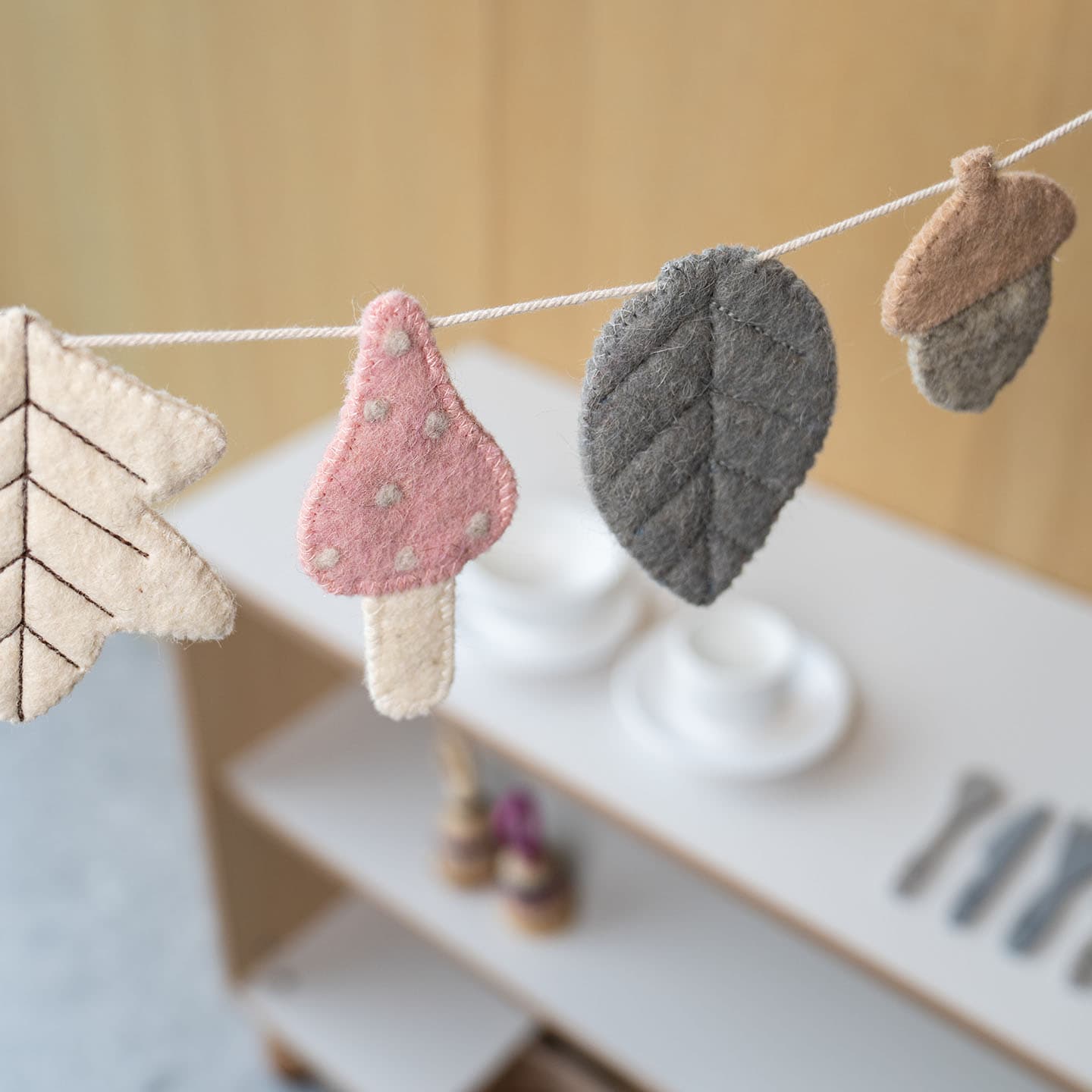 Een slinger met vilten vormen van een blad, paddenstoel en eikel hangt in een minimalistische kamer, waar borden en bestek wazig op de achtergrond te zien zijn.