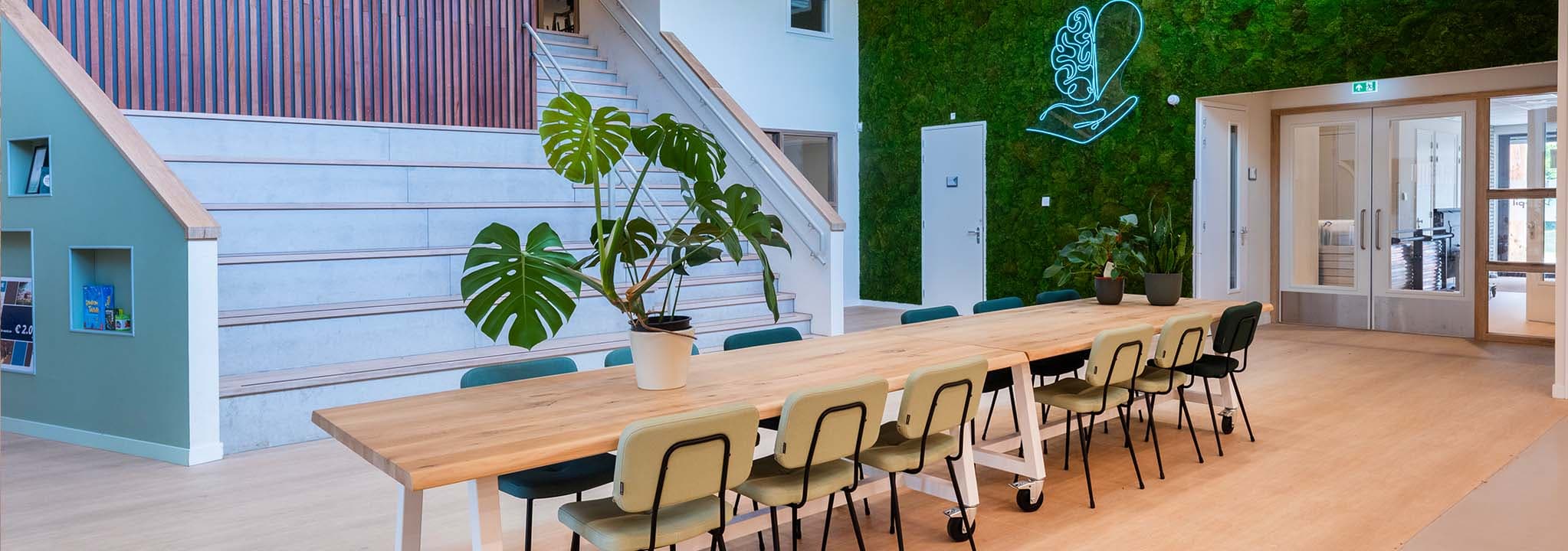 Centrale groepsruimte in IKC De Spil met veel zitplekken aan verrijdbare grote houten tafels en een groene plantenwand.