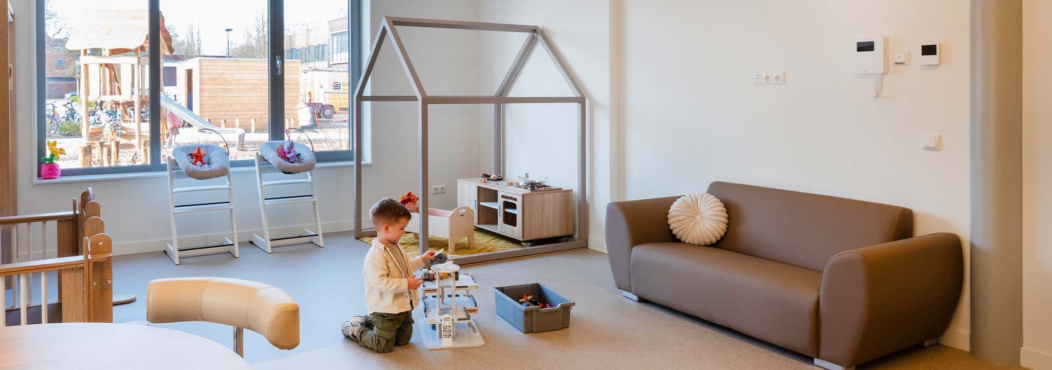 Een kind speelt met speelgoed in een moderne, ruime speelkamer met een bank, speelgoedkeuken en een groot raam dat zorgt voor natuurlijk licht.