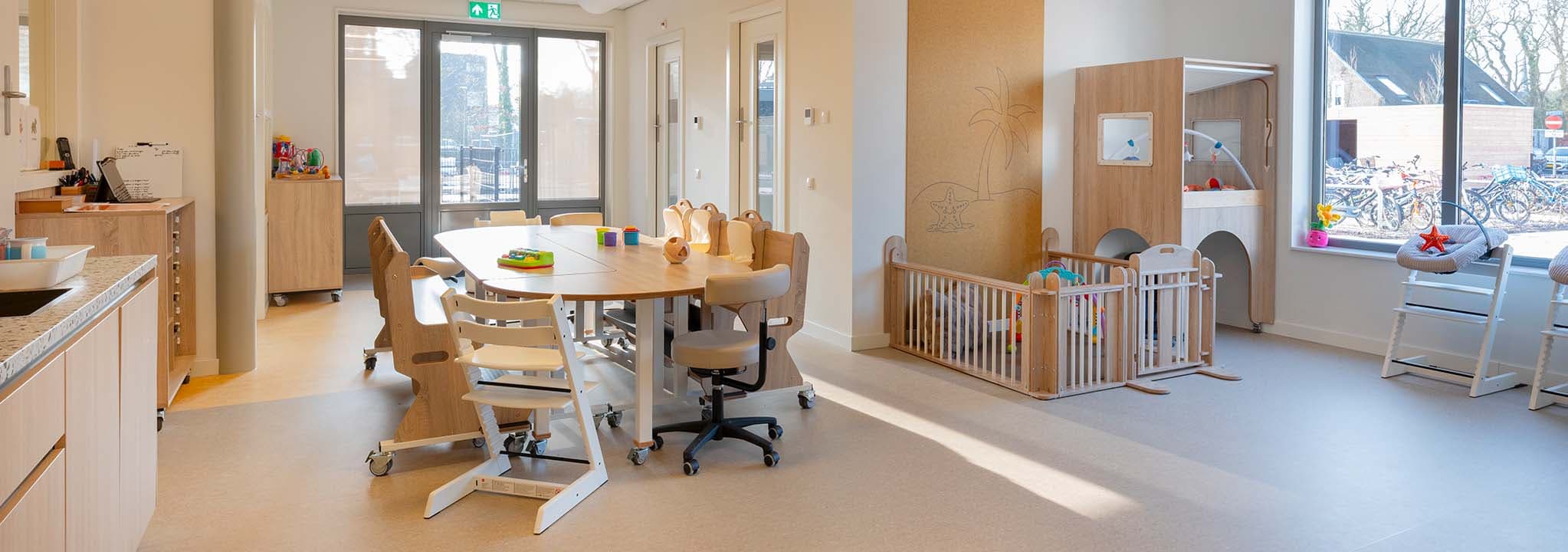 Lichte groepsruimte voor kinderen van 0 tot 2 jaar met een ovale verrijdbare houten kindertafel, ergobanken, kinderstoelen, een houten grondbox en een verhoogde box met speelhoek eronder. Grote ramen zorgen voor veel daglicht.