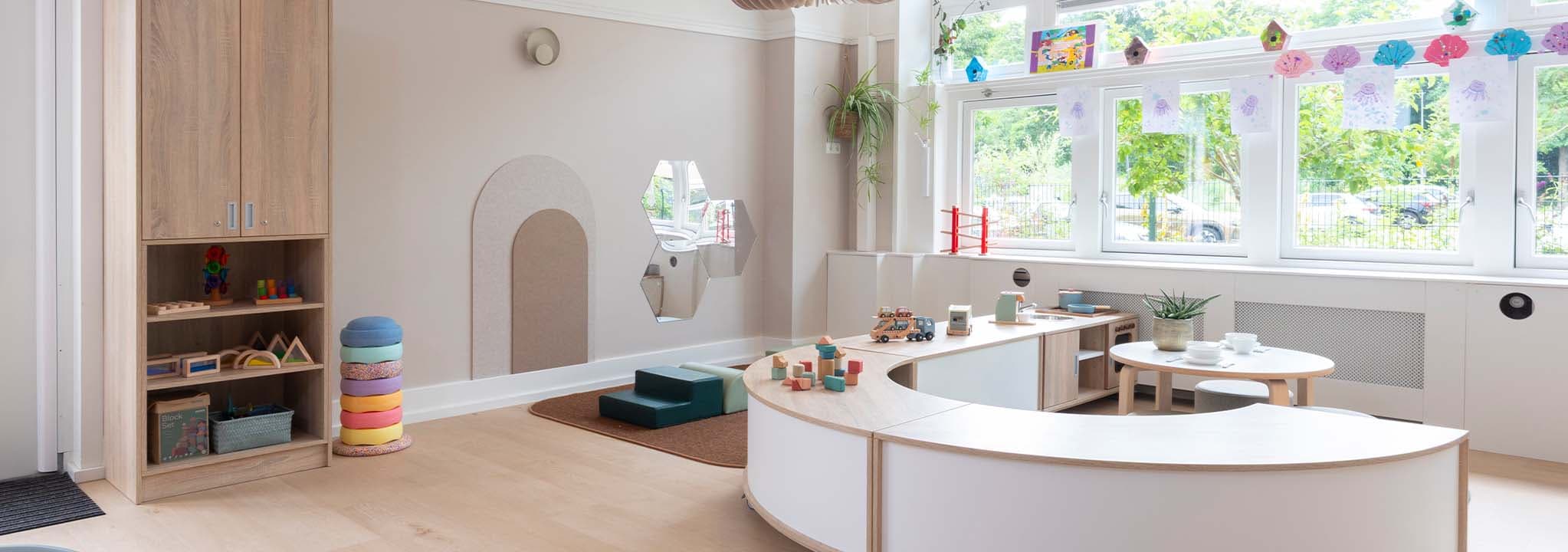 Een lichte, ruime groepsruimte in een kinderdagverblijf met houten meubels, kleurrijk speelgoed en grote ramen die zijn versierd met knutselwerkjes.