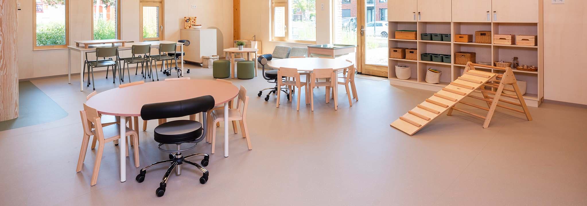 Ruime en lichte groepsruimte in de kinderopvang met ronde houten tafels, stoelen, opbergkasten tegen de muur, een klimdriehoek en grote ramen met veel daglicht.