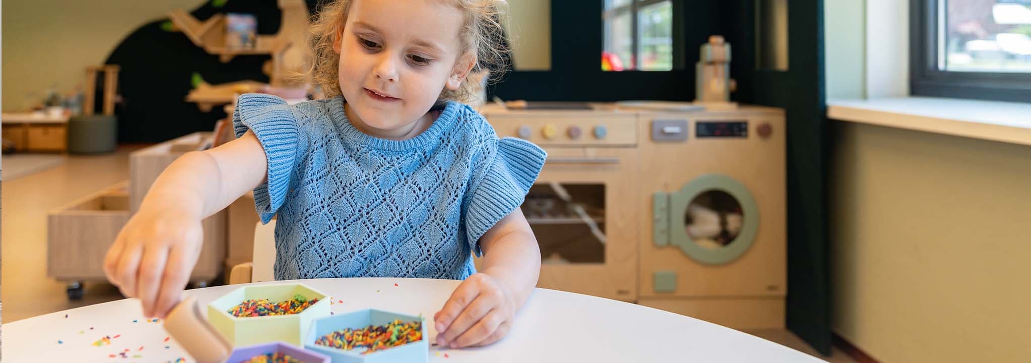 Een kind in een blauwe trui speelt met kleurrijke stukken aan een tafel in een speelkamer, met speelgoedkeukenapparatuur op de achtergrond.