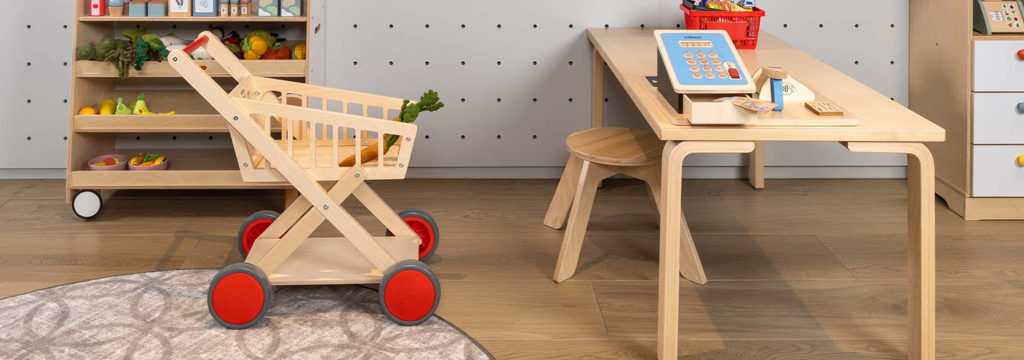 Kinderspeelkamer met een houten tafel, stoel, speelgoedwinkelwagen, speelgoedeten op planken en een kleurrijke mand met bouwblokken op de tafel.