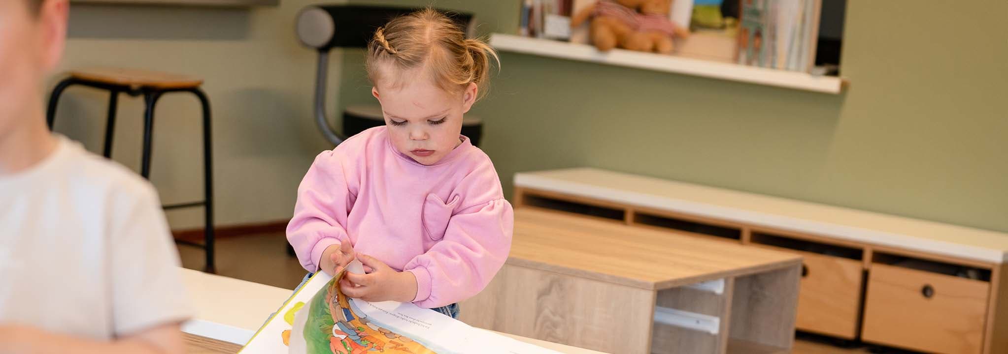Een jong kind in een roze trui kijkt naar een boek in een gezellige kamer met boekenplanken en een krukje op de achtergrond.