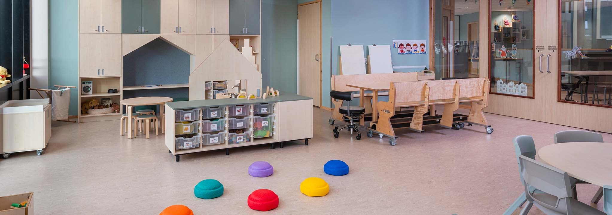 Een lichte groepsruimte in een kinderdagverblijf met kleurrijke kussens, houten meubels, planken en kasten. Tafels en stoelen staan netjes opgesteld tegen een lichtblauwe muur.