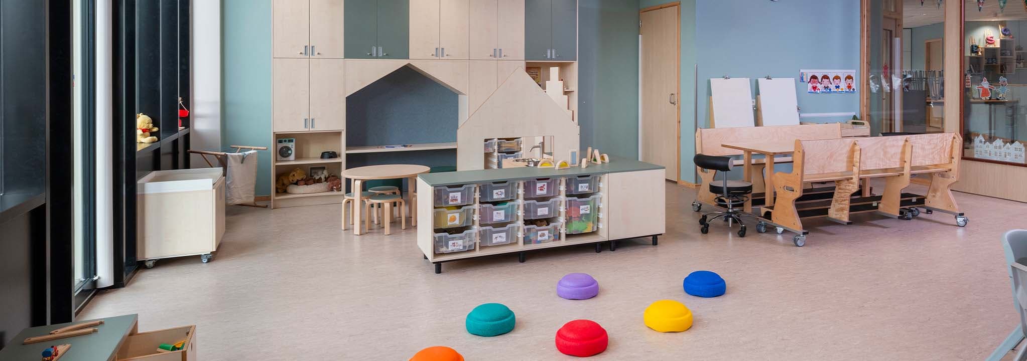 Groepsruimte in de kinderopvang met verrijdbaar meubilair, waaronder een houten tafel, houten banken, lage opbergkasten en een op maat gemaakte houten kast.