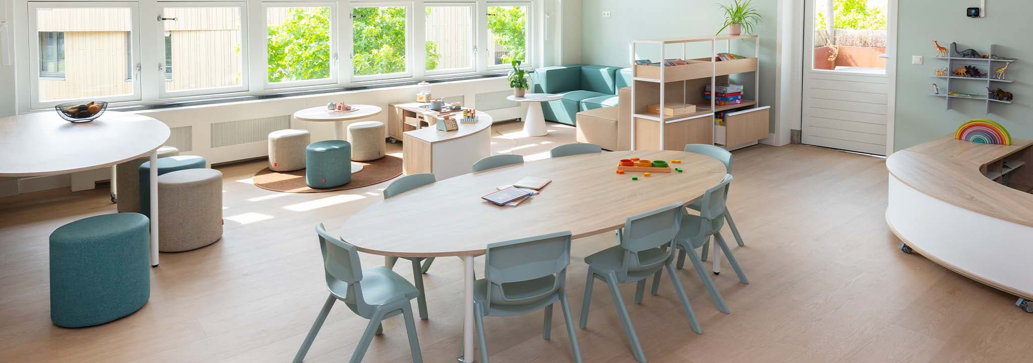 Lichte groepsruimte in de kinderopvang met grote ramen, een lage ovale houten tafels met lichtgroene kinderstoelen, daarachter een knusse bank en andere zithoeken en open opbergkasten.