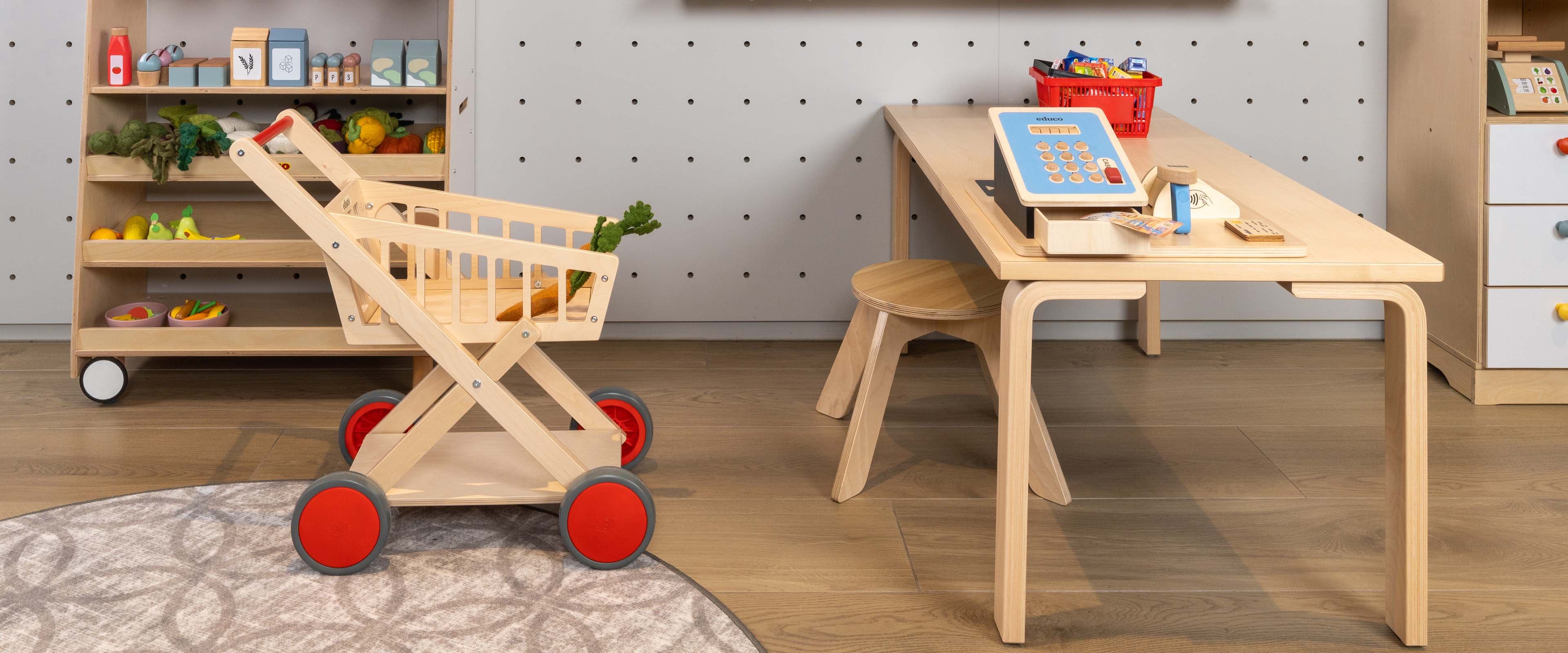 Kinderspeelkamer met een houten tafel, stoel, speelgoedwinkelwagen, speelgoedeten op planken en een kleurrijke mand met bouwblokken op de tafel.