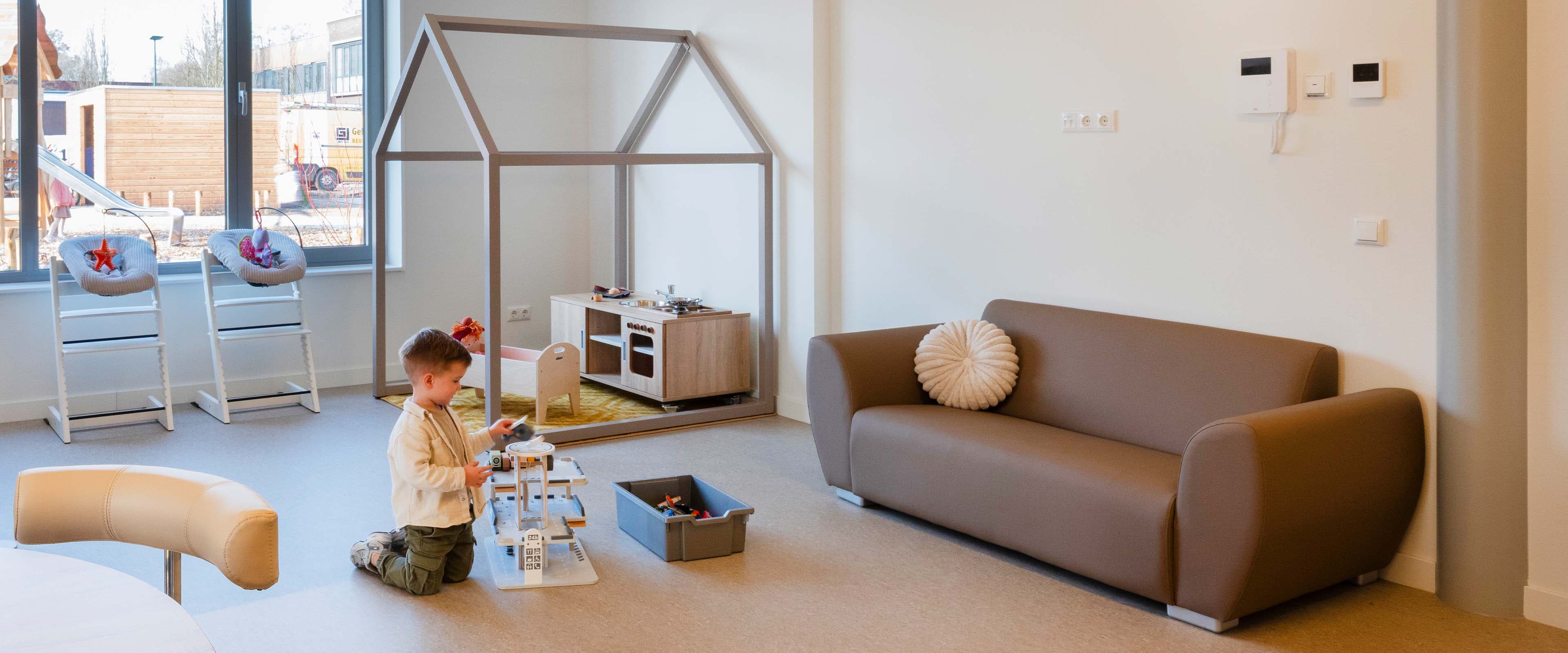 Een kind speelt met speelgoed in een moderne, ruime speelkamer met een bank, speelgoedkeuken en een groot raam dat zorgt voor natuurlijk licht.