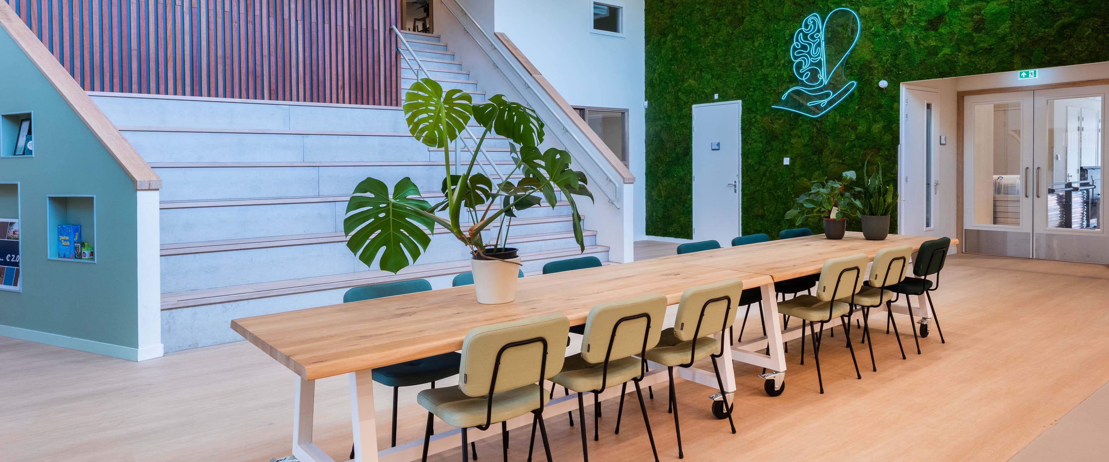 Centrale groepsruimte in IKC De Spil met veel zitplekken aan verrijdbare grote houten tafels en een groene plantenwand.