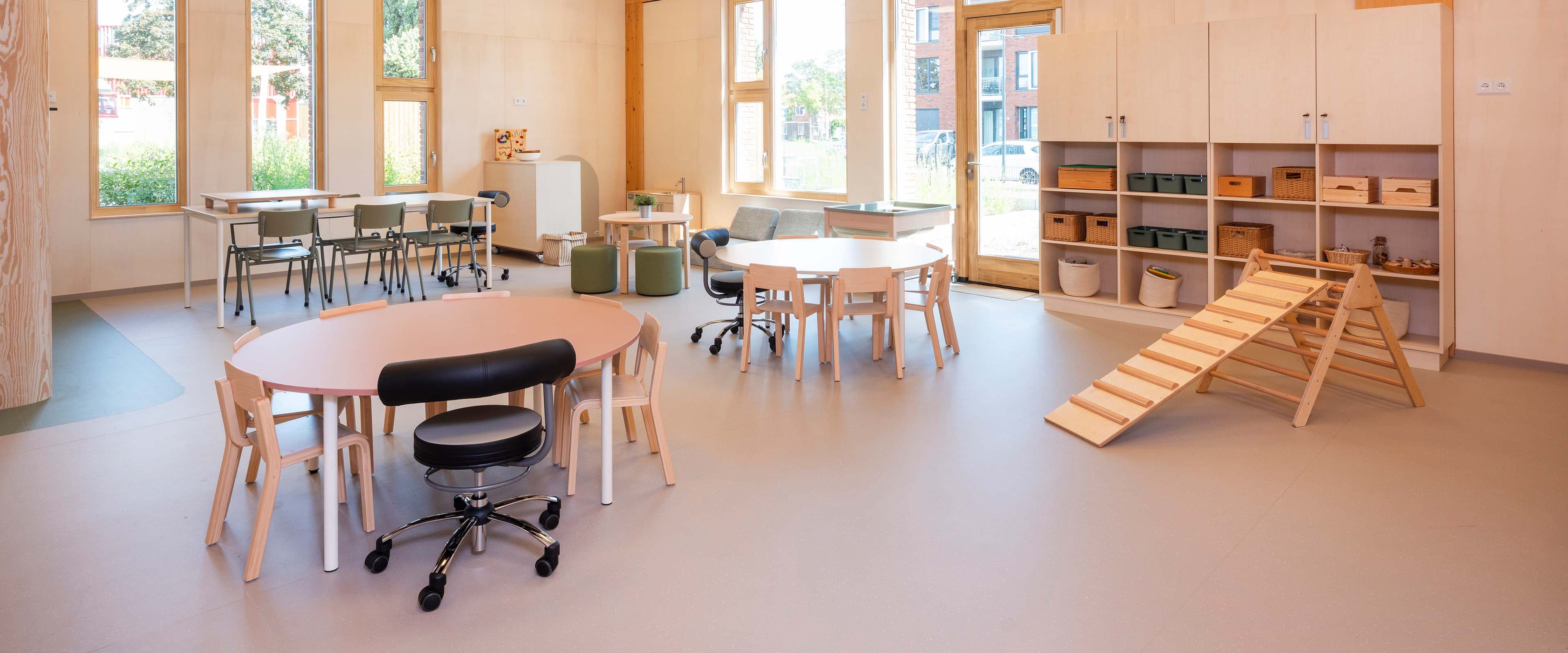 Ruime en lichte groepsruimte in de kinderopvang met ronde houten tafels, stoelen, opbergkasten tegen de muur, een klimdriehoek en grote ramen met veel daglicht.