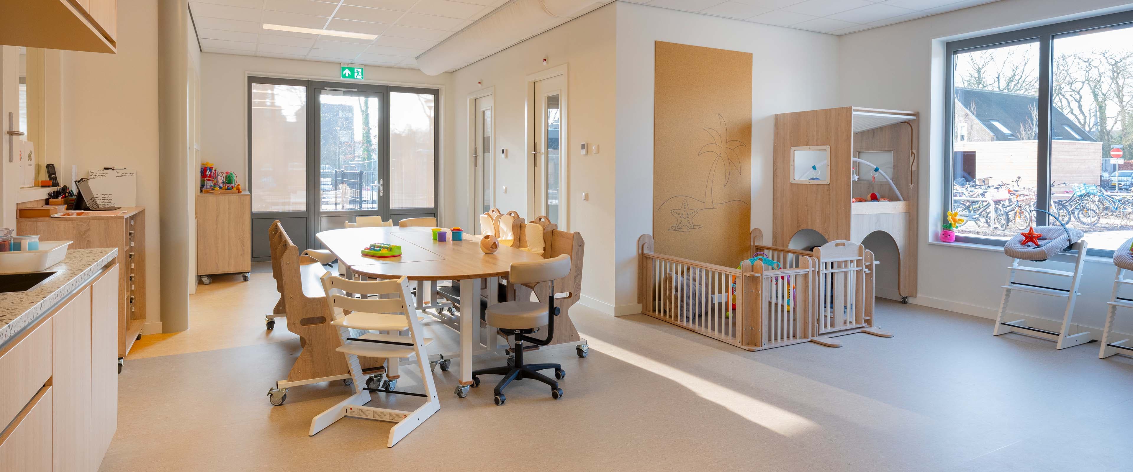 Lichte groepsruimte voor kinderen van 0 tot 2 jaar met een ovale verrijdbare houten kindertafel, ergobanken, kinderstoelen, een houten grondbox en een verhoogde box met speelhoek eronder. Grote ramen zorgen voor veel daglicht.