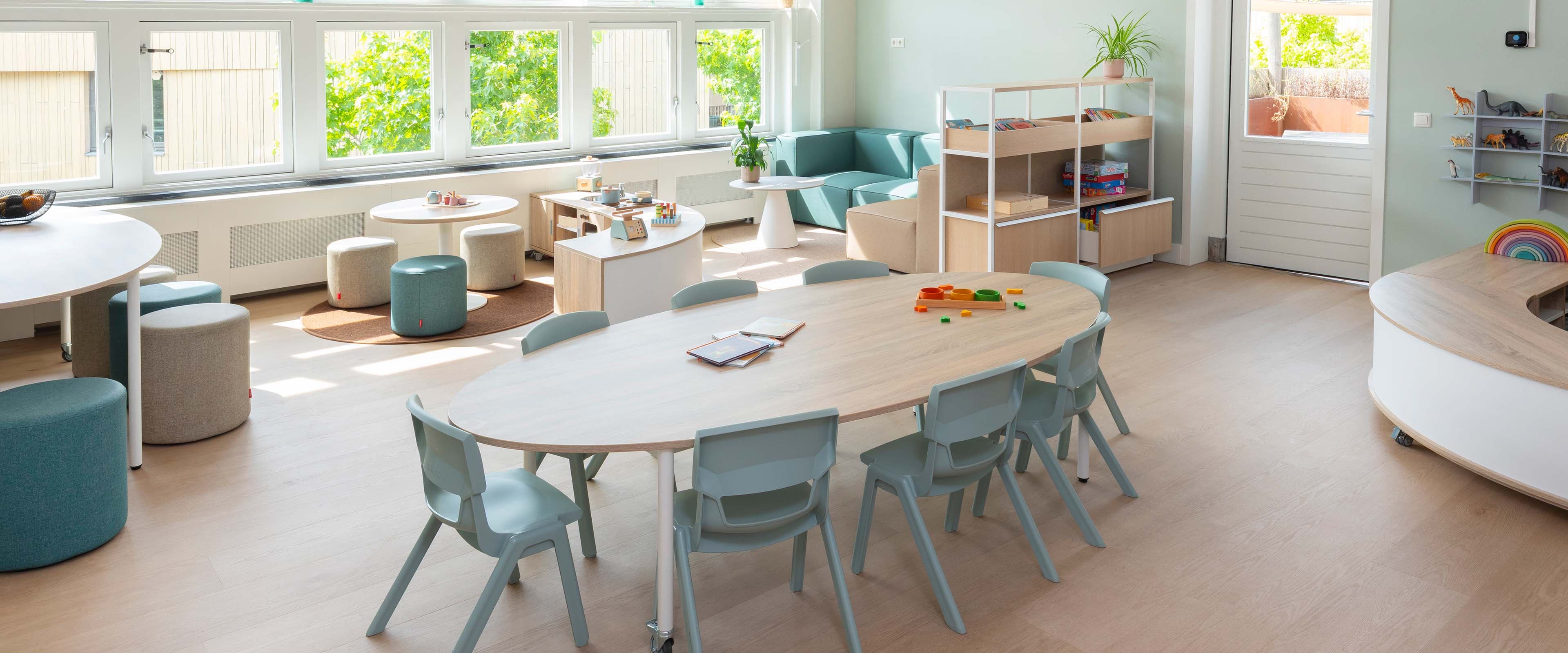 Lichte groepsruimte in de kinderopvang met grote ramen, een lage ovale houten tafels met lichtgroene kinderstoelen, daarachter een knusse bank en andere zithoeken en open opbergkasten.