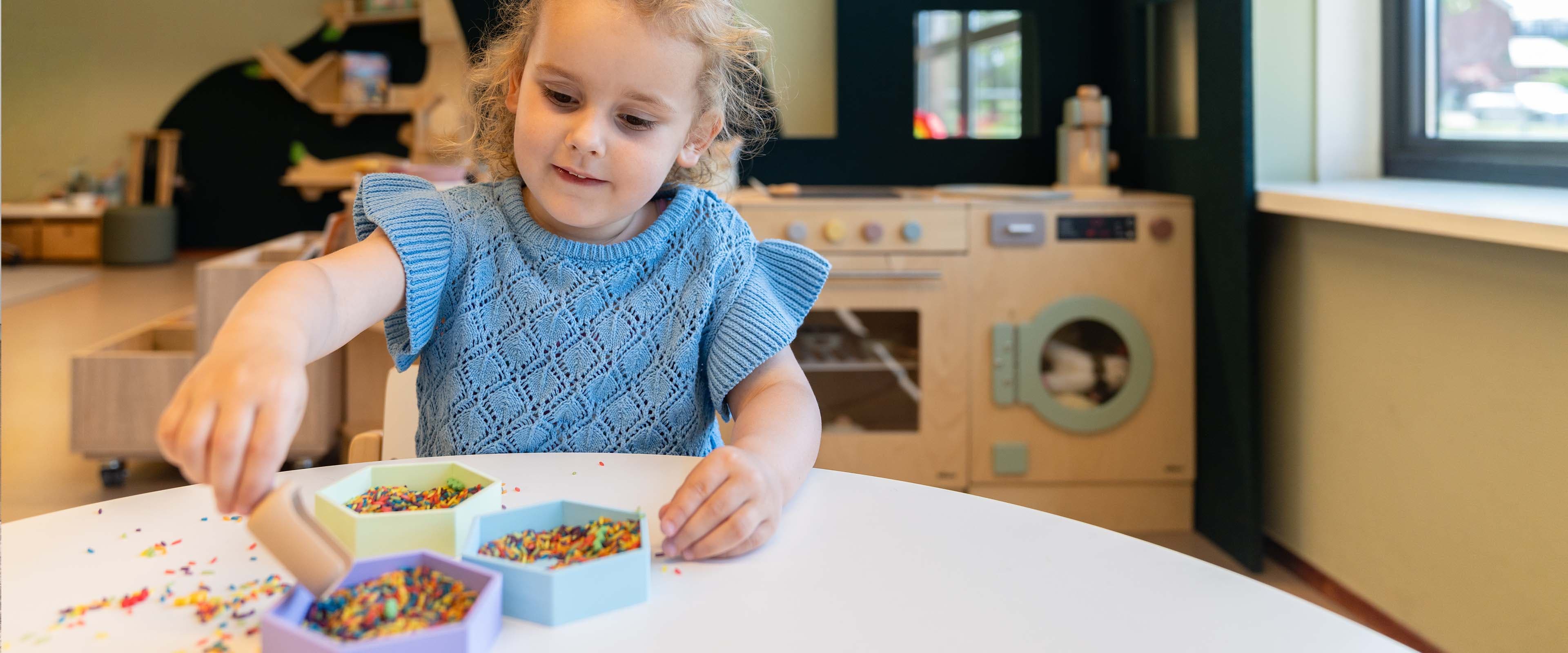 Een kind in een blauwe trui speelt met kleurrijke stukken aan een tafel in een speelkamer, met speelgoedkeukenapparatuur op de achtergrond.