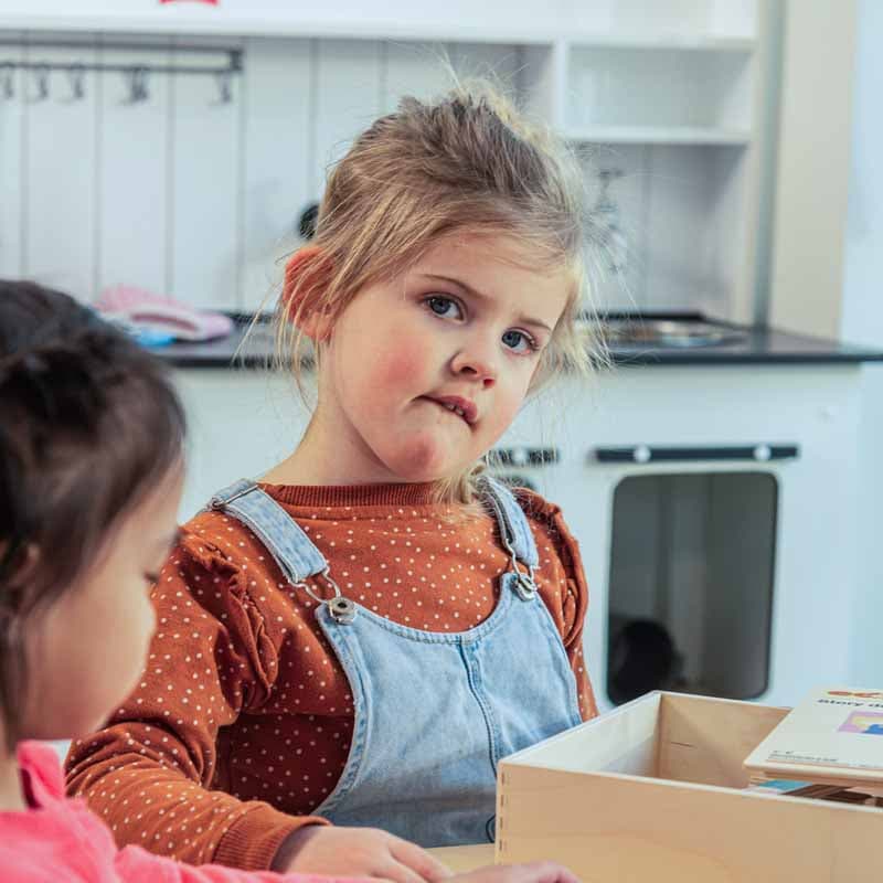 Twee jonge meisjes spelen in een keukentje. Een van de meisjes, in een shirt met stippen en een spijkeroverall, kijkt nieuwsgierig in een houten doos.