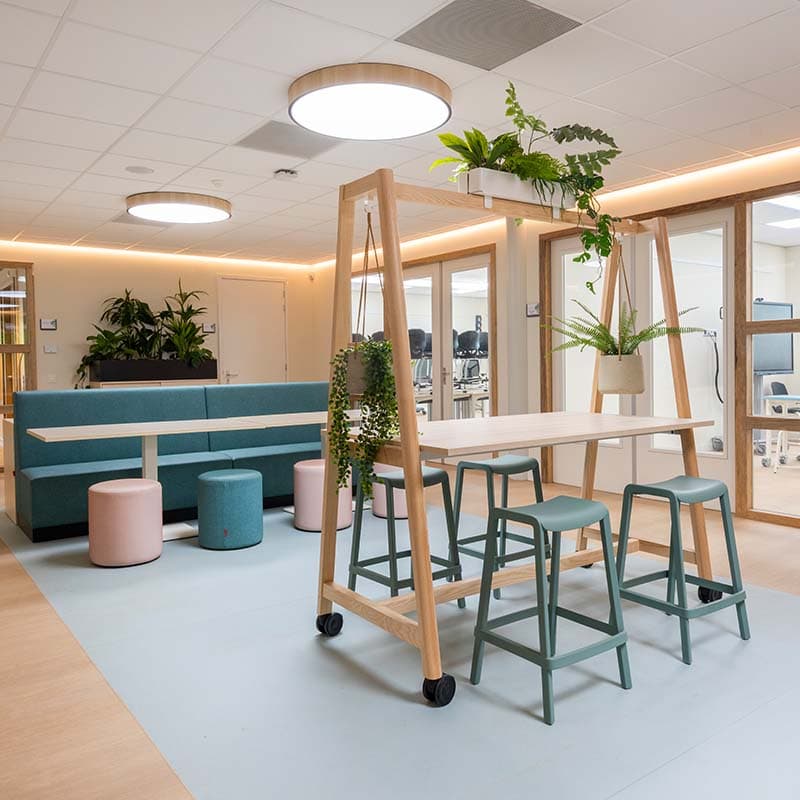 Open ruimte in de kinderopvang met een verrijdbare houten tafel, krukken, zitbanken en hangplanten in een lichte omgeving.