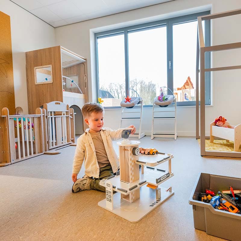 Een dreumes speelt met een houten parkeergarage in een lichte, gezellige speelruimte van een kinderdagverblijf. Op de achtergrond zijn een bedje en speelgoed zichtbaar.