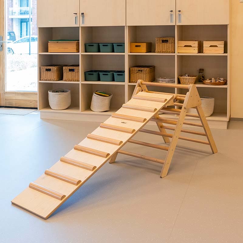 Houten klimtoestel met hellingbaan in een groepsruimte met houten open opbergkasten waarin opbergbakken geplaatst zijn.