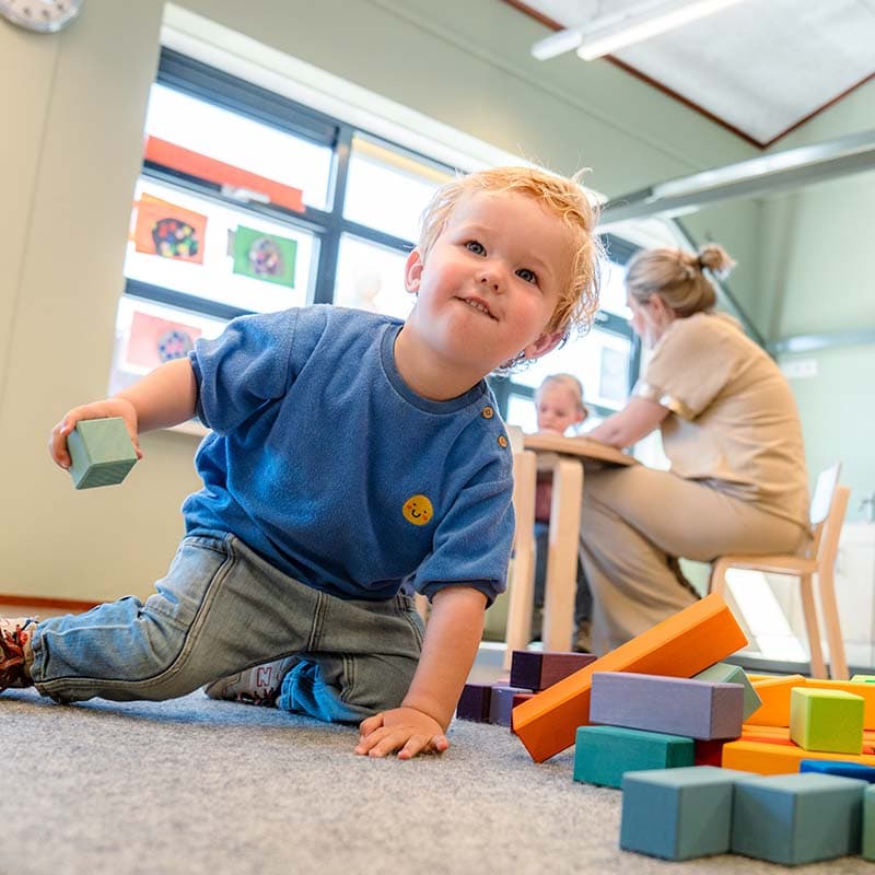 Een jong kind in een blauw shirt speelt met kleurrijke blokken op de grond, terwijl een pedagogisch professional en een ander kind op de achtergrond aan een tafel zitten.