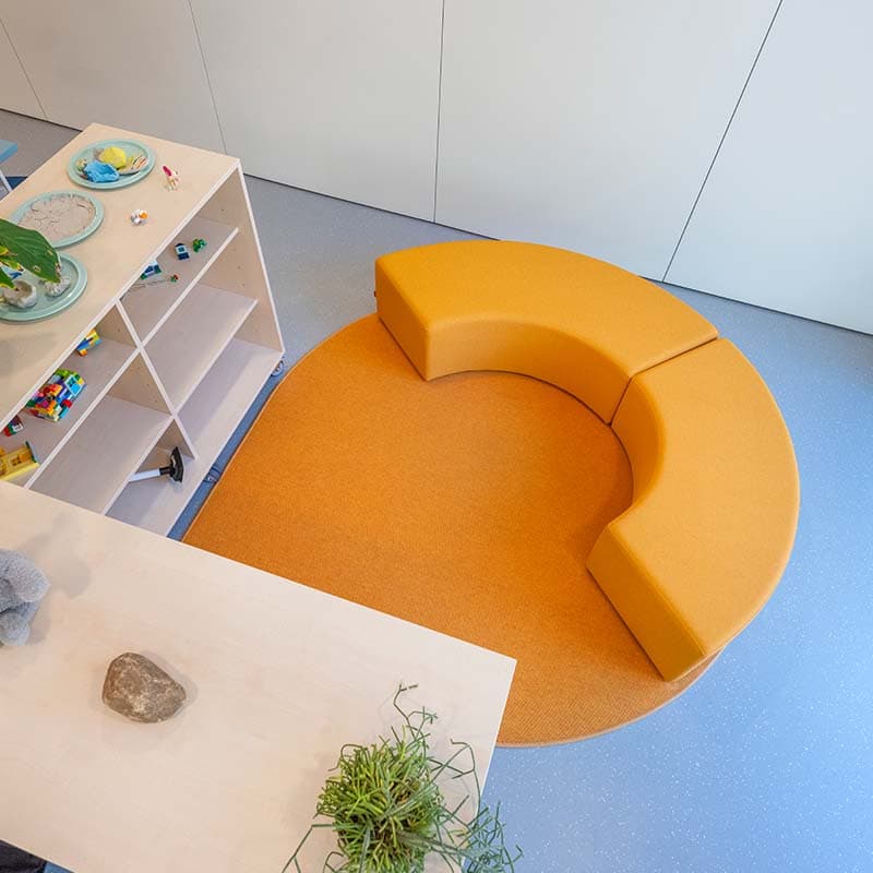 Gebogen oranje zitbank en vloerkleed in een speelhoek met opbergkasten, spelmateriaal en planten.