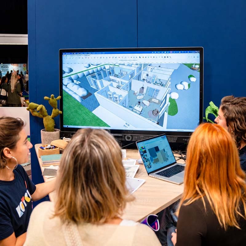 Vier mensen werken samen aan een bureau en bekijken een 3D-model van een gebouw op een groot scherm, met een laptop waarop ontwerpopties worden getoond.