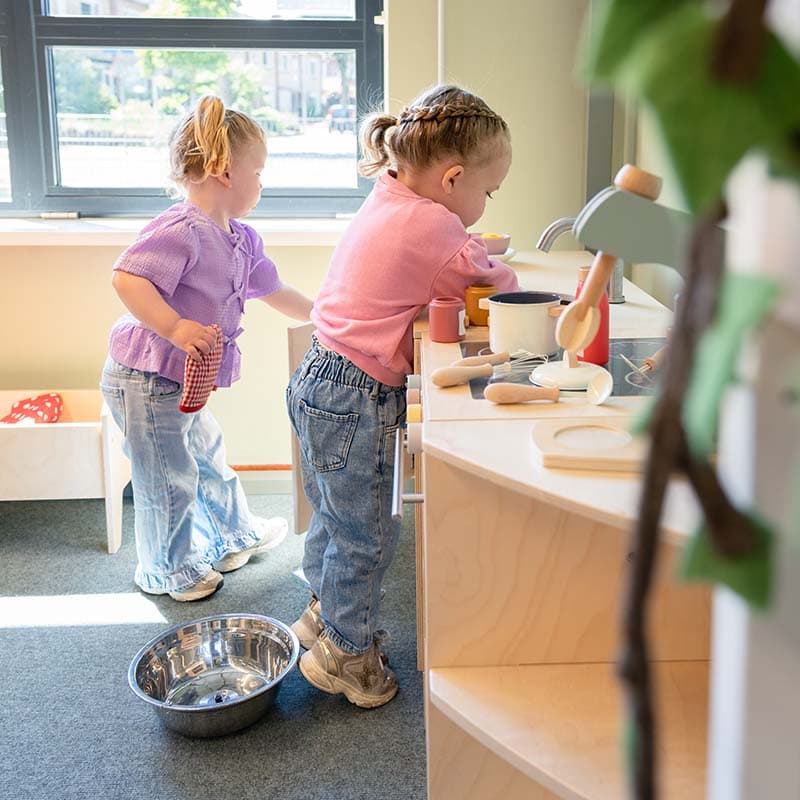 Twee kinderen spelen in een speelkeuken; de ene roert in een pan, de andere houdt een doek vast. Zonlicht stroomt naar binnen door een groot raam.