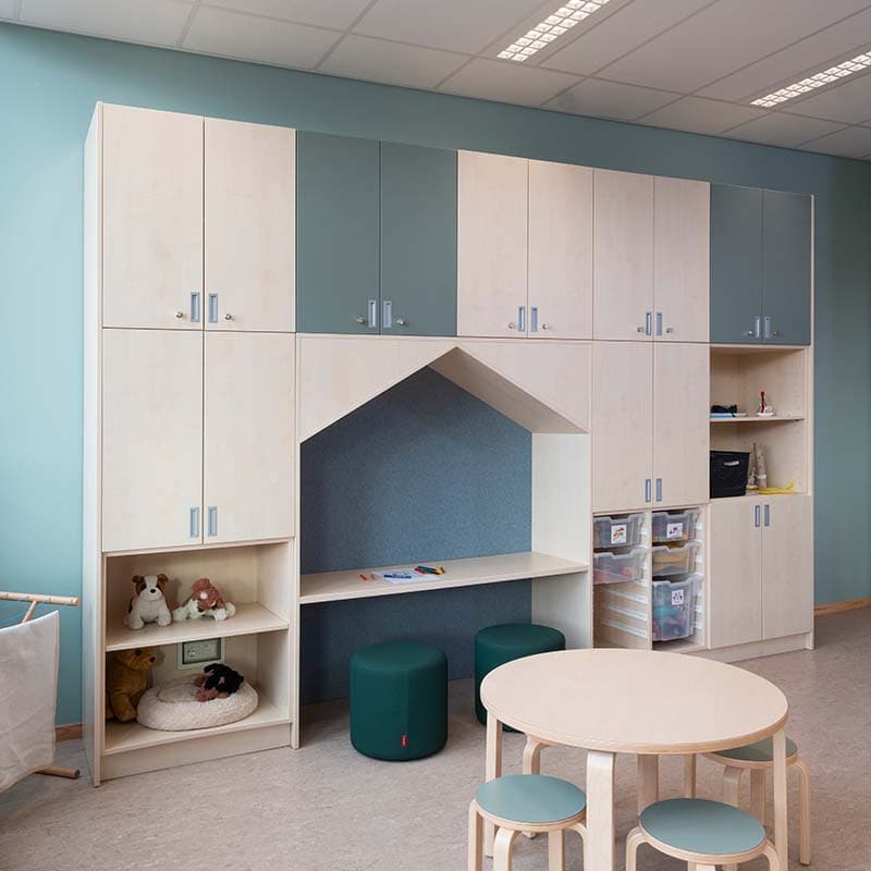 Op maat gemaakte houten wandkast in de kinderopvang met een ingebouwd speelhuisje, opbergruimte en poefjes onder een werkblad.