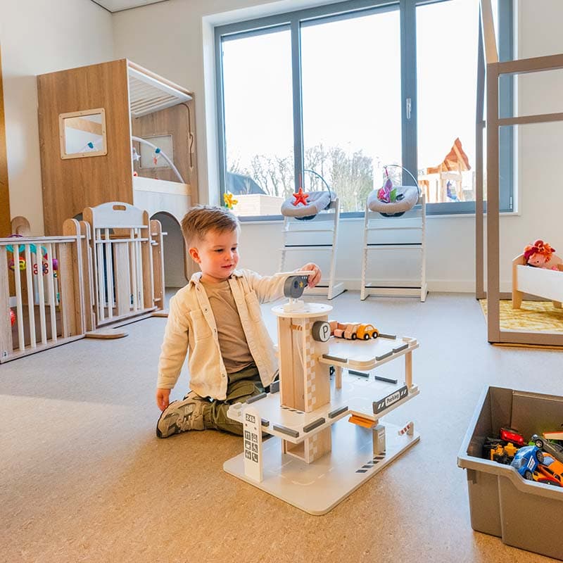 Kind speelt met een speelgoedgarage op de vloer in een lichte groepsruimte met spelmateriaal en grote ramen.